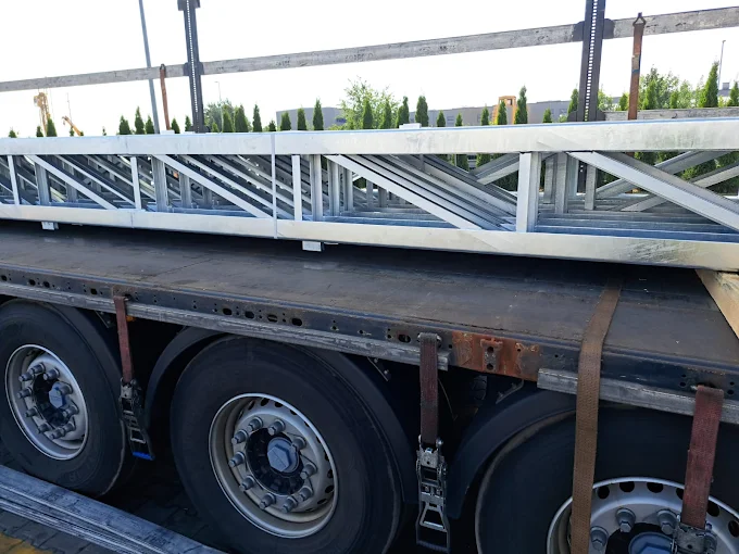 Metalowe elementy konstrukcyjne przewożone na naczepie ciężarówki, zabezpieczone pasami transportowymi