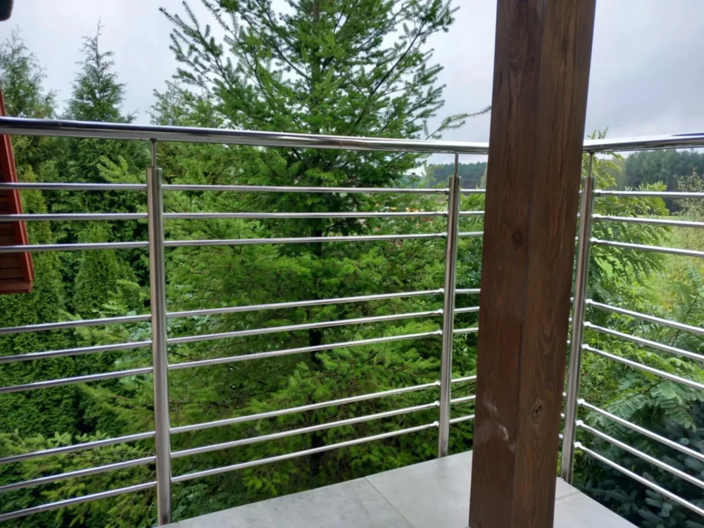 Widok z balkonu z metalową balustradą ze stali nierdzewnej i drewnianym słupem, w tle gęste zielone drzewa i las.