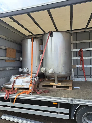 Dwa duże stalowe zbiorniki ustawione na paletach i zabezpieczone pasami transportowymi wewnątrz ciężarówki.