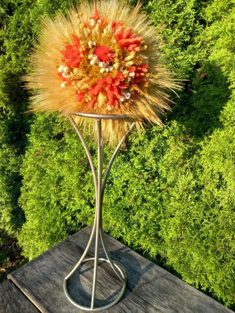 Bukiet z suszonych zbóż i kwiatów w odcieniach złota, czerwieni i bieli, umieszczony na wysokim, metalowym stojaku, ustawiony na tle zielonych krzewów