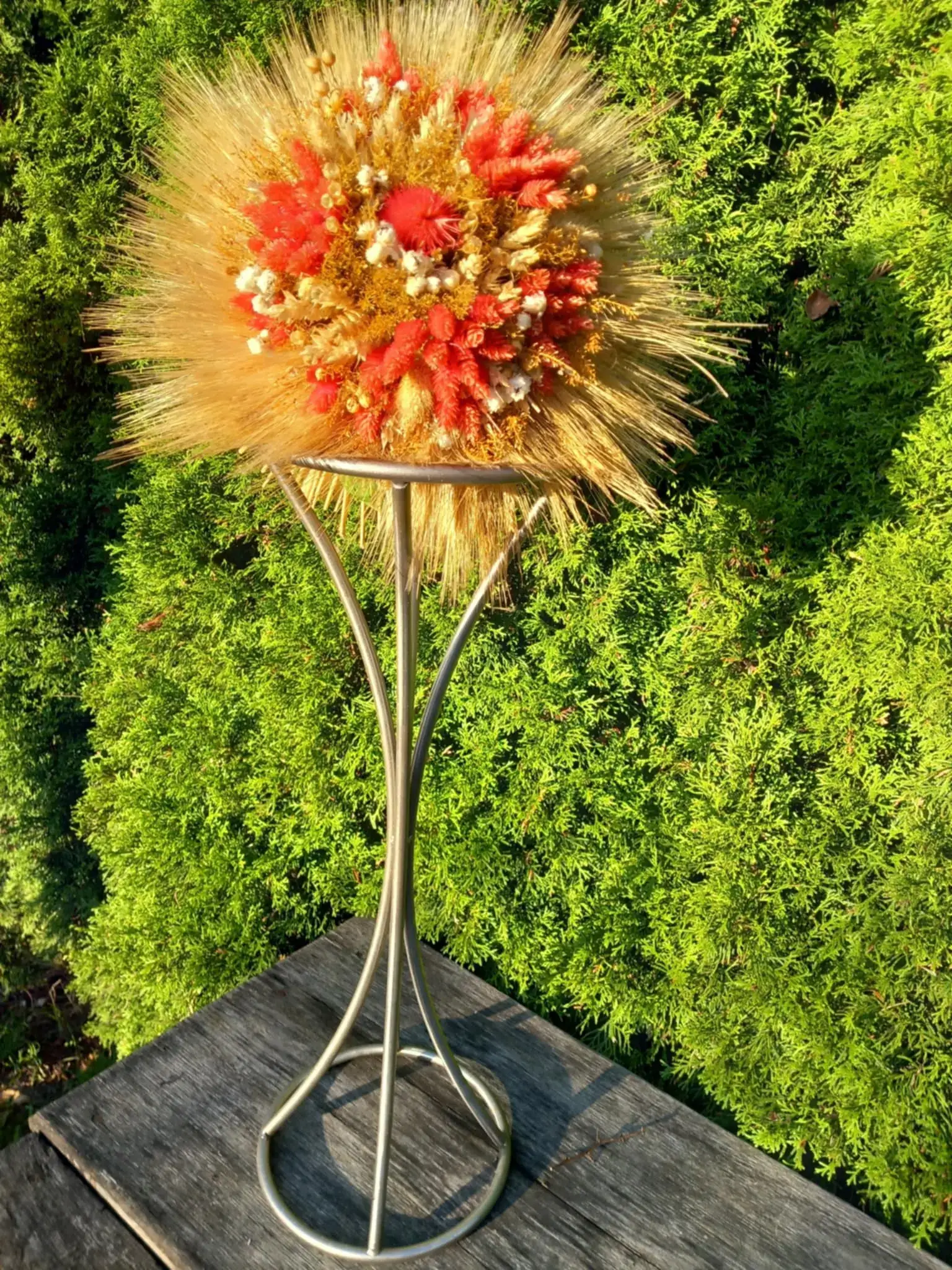 Bukiet z suszonych zbóż i kwiatów w odcieniach złota, czerwieni i bieli, umieszczony na wysokim, metalowym stojaku, ustawiony na tle zielonych krzewów