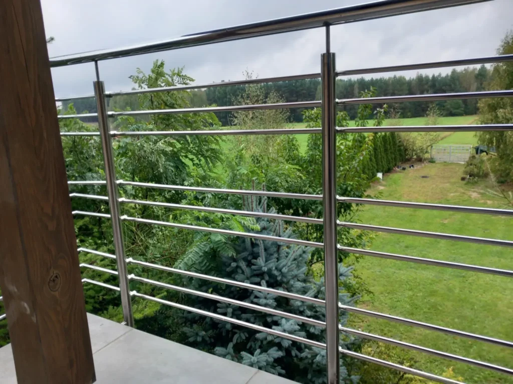 Widok z balkonu z metalową balustradą ze stali nierdzewnej i drewnianym słupem, w tle gęste zielone drzewa i las.