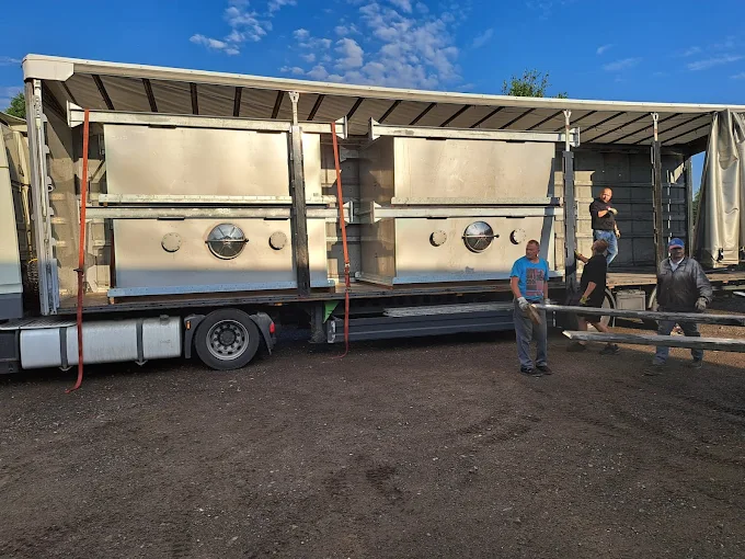 Załadunek dużych metalowych zbiorników na naczepę ciężarową, kilku pracowników pomaga przy transporcie na otwartym placu.