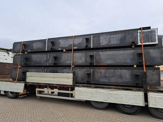 Ciężarówka z otwartą naczepą przewożąca duże stalowe belki konstrukcyjne, zabezpieczone pasami transportowymi.