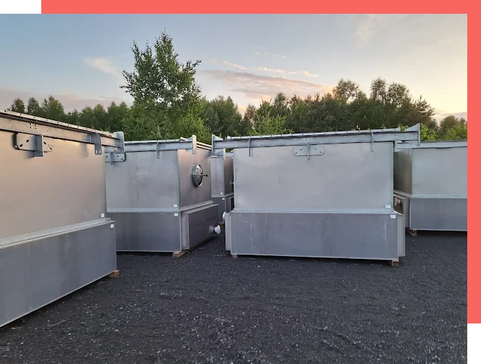 Metalowe kontenery przemysłowe ustawione na placu z żwiru, w tle drzewa i zachód słońca