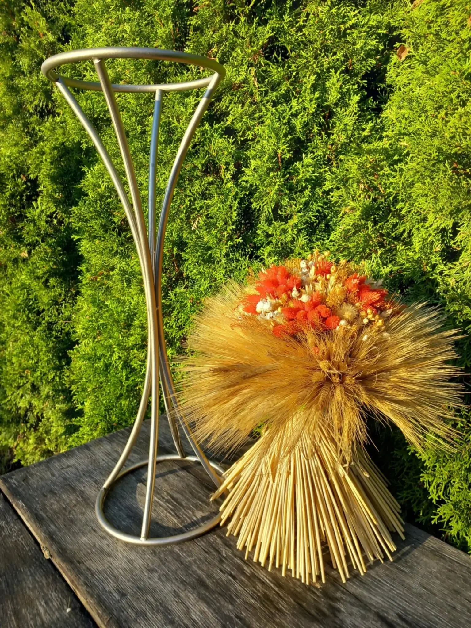 Bukiet z suszonych zbóż i kwiatów w odcieniach złota, czerwieni i bieli, umieszczony na wysokim, metalowym stojaku, ustawiony na tle zielonych krzewów