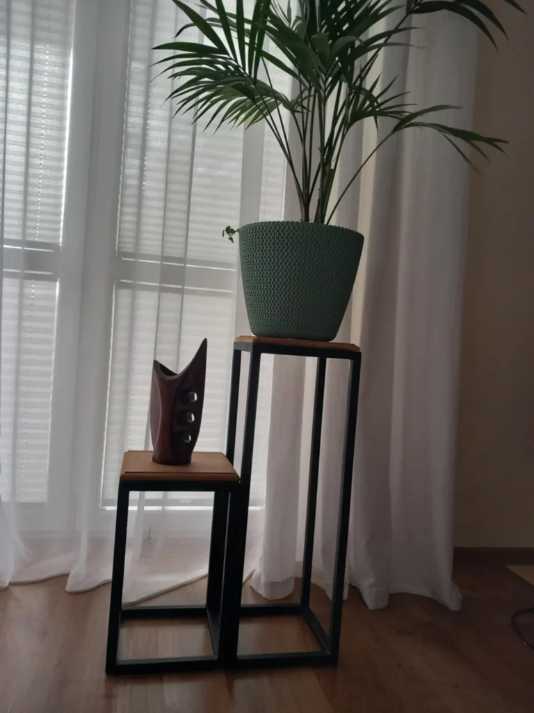 Nowoczesne stojaki w formie metalowych ramek z drewnianymi blatami. Na niższym stoi drewniana dekoracja o organicznych kształtach z trzema okrągłymi otworami, a na wyższym znajduje się zielona doniczka z rośliną. Tło stanowią białe zasłony i okno z żaluzjami.