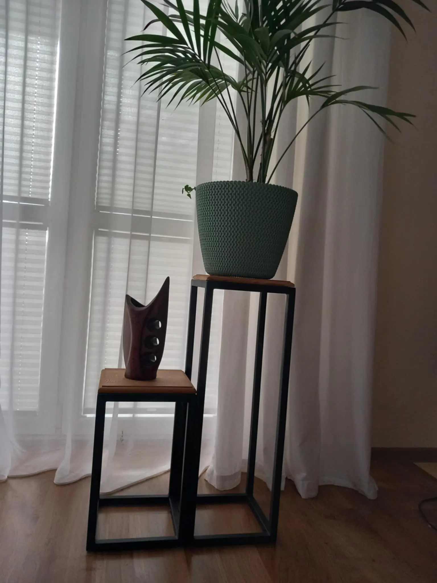 Nowoczesne stojaki w formie metalowych ramek z drewnianymi blatami. Na niższym stoi drewniana dekoracja o organicznych kształtach z trzema okrągłymi otworami, a na wyższym znajduje się zielona doniczka z rośliną. Tło stanowią białe zasłony i okno z żaluzjami.