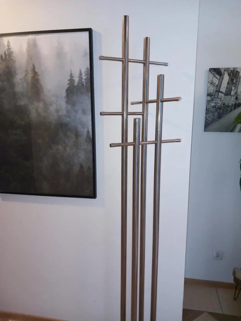 Nowoczesny metalowy wieszak stojący z kilkoma poziomymi uchwytami na tle białej ściany, obok którego wiszą dwa obrazy – po lewej z motywem mglistych drzew, a po prawej z miejską scenerią.