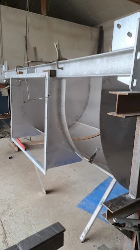 Metalowa konstrukcja spawana w warsztacie, składająca się z dużych zakrzywionych blach i stalowych profili, ustawiona na stojakach.