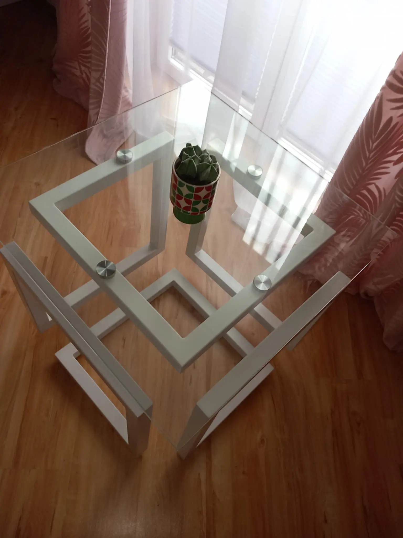 Szklany stolik z białą, geometryczną ramą stojący na drewnianej podłodze, na środku blatu w kolorowej doniczce znajduje się mały kaktus; w tle różowe zasłony i białe firanki przepuszczające światło dzienne.