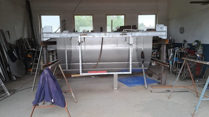 Warsztat metalowy z dużą, niedokończoną konstrukcją aluminiową ustawioną na stojakach, otoczony narzędziami i sprzętem