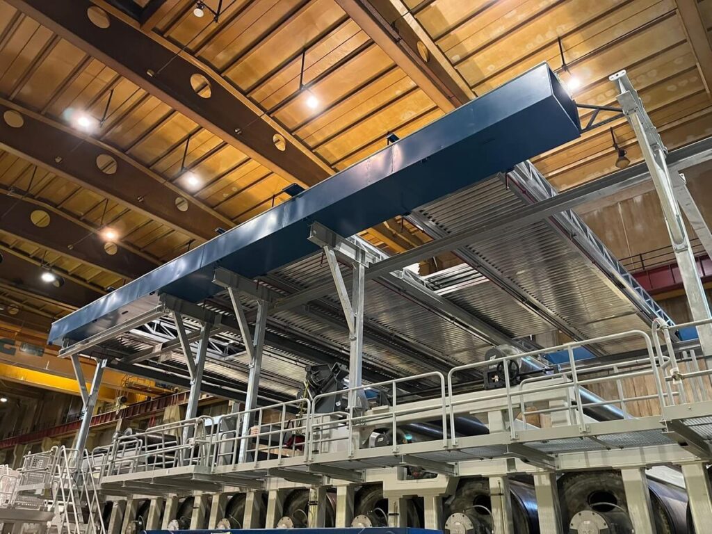 Stalowa konstrukcja przemysłowa wewnątrz hali produkcyjnej, z platformą roboczą, rurami i instalacjami technicznymi pod sufitem.