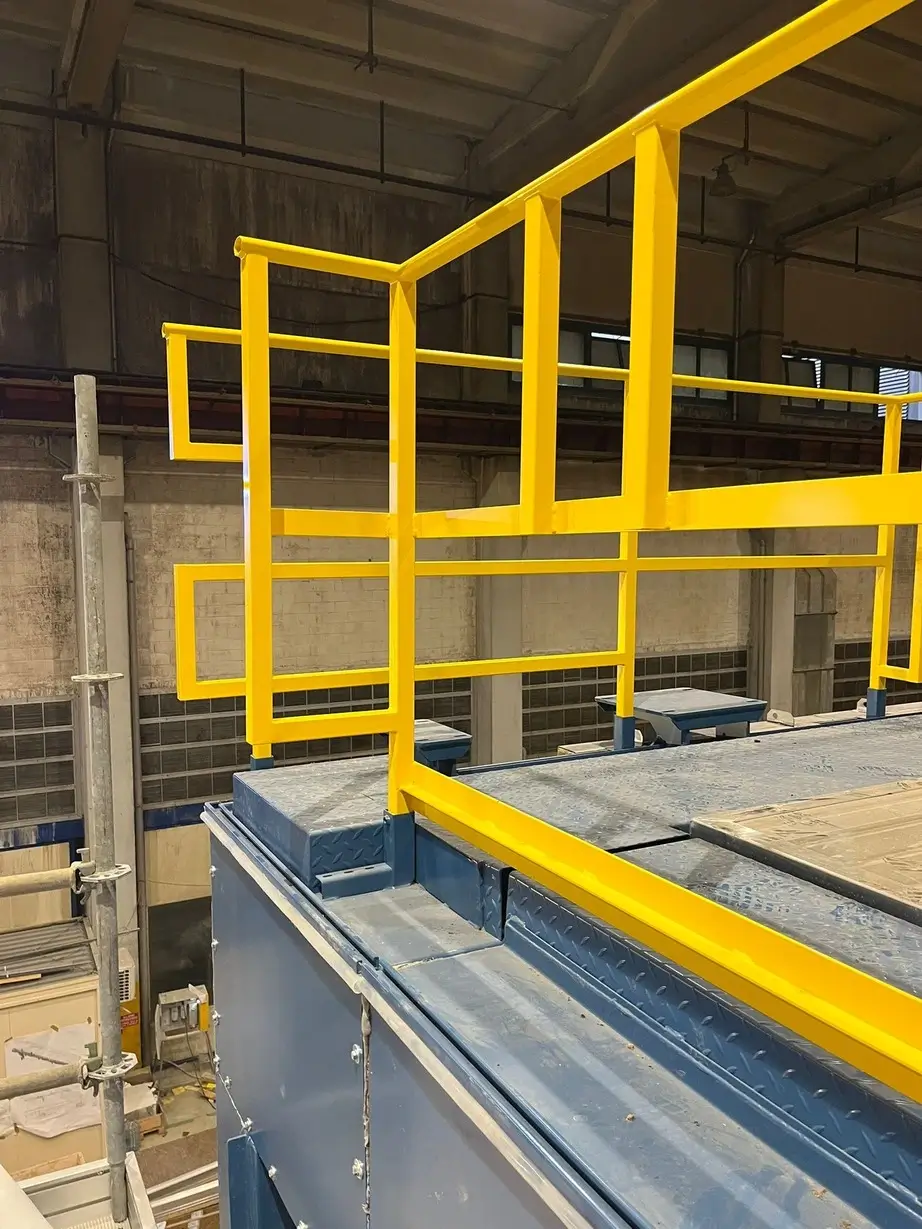 Barierka ochronna w kolorze żółtym zamontowana na metalowej konstrukcji wewnątrz hali przemysłowej.