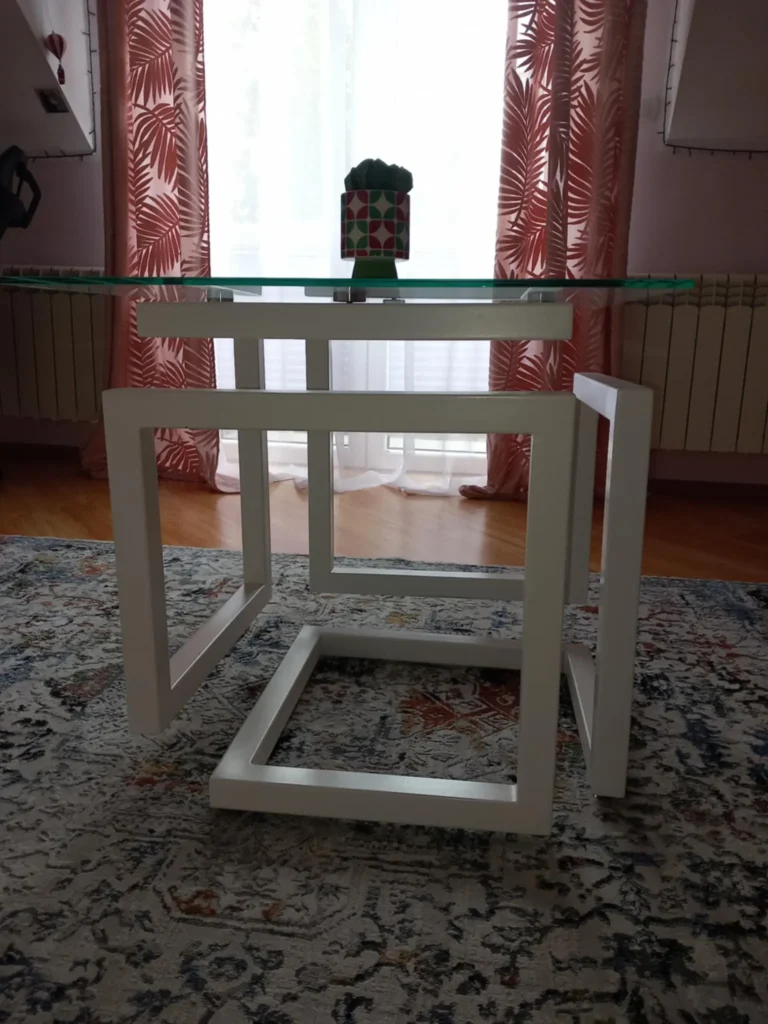 Szklany stolik z białą, geometryczną ramą stojący na drewnianej podłodze, na środku blatu w kolorowej doniczce znajduje się mały kaktus; w tle różowe zasłony i białe firanki przepuszczające światło dzienne.