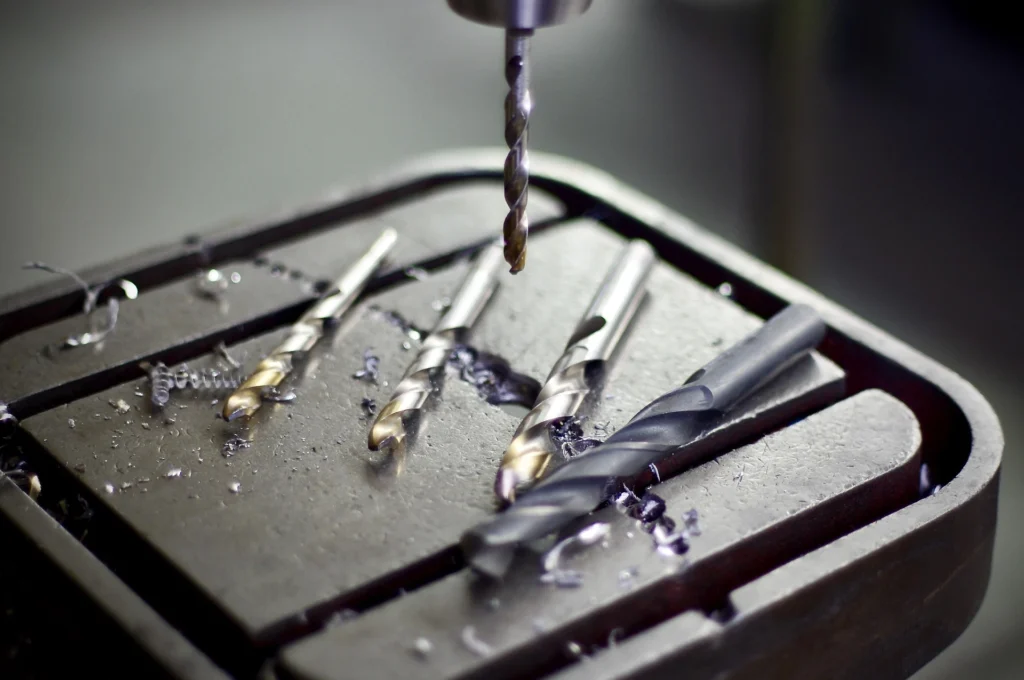 Wiertło wiercące otwór w metalowym bloku. Obok leżą metalowe opiłki i inne zużyte już wiertła.