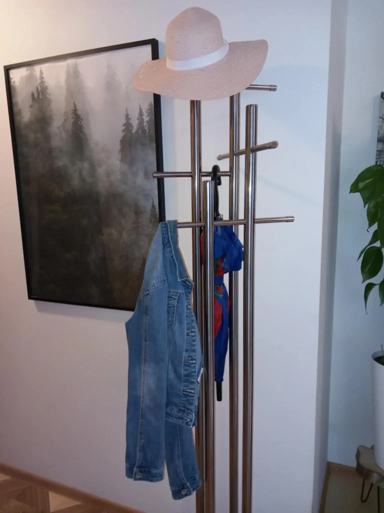 Metalowy wieszak stojący w przedpokoju z zawieszonym kapeluszem, dżinsową kurtką i kolorową parasolką, obok obrazu przedstawiającego las we mgle.