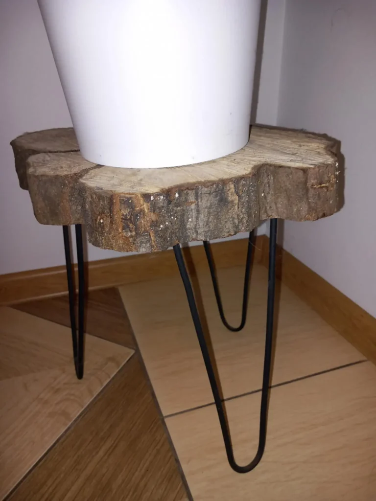 Mały stolik lub stojak z blatem z naturalnego, nieobrobionego plastra drewna na czarnych metalowych nóżkach, na którym stoi biały okrągły doniczkowy pojemnik, ustawiony na styku drewnianej i kafelkowej podłogi w rogu pomieszczenia.